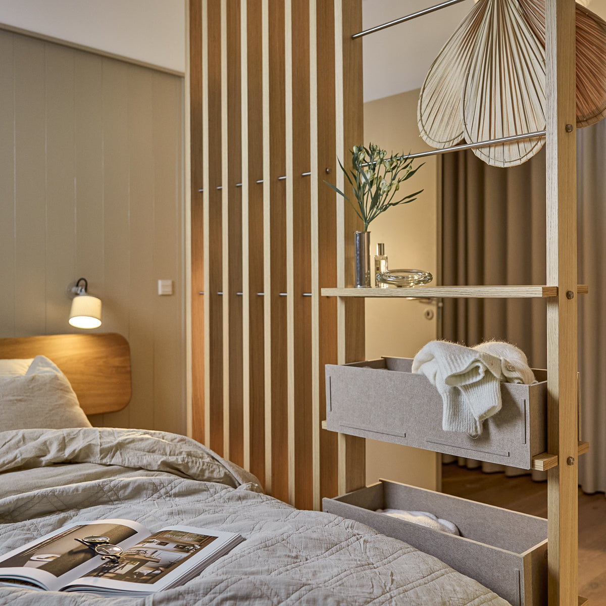 Une chambre à coucher moderne avec une literie nordique, une lampe suspendue et un magazine sur le lit soigneusement fait. Des panneaux acoustiques en bois Akupanel de WoodUpp rehaussent le mur.